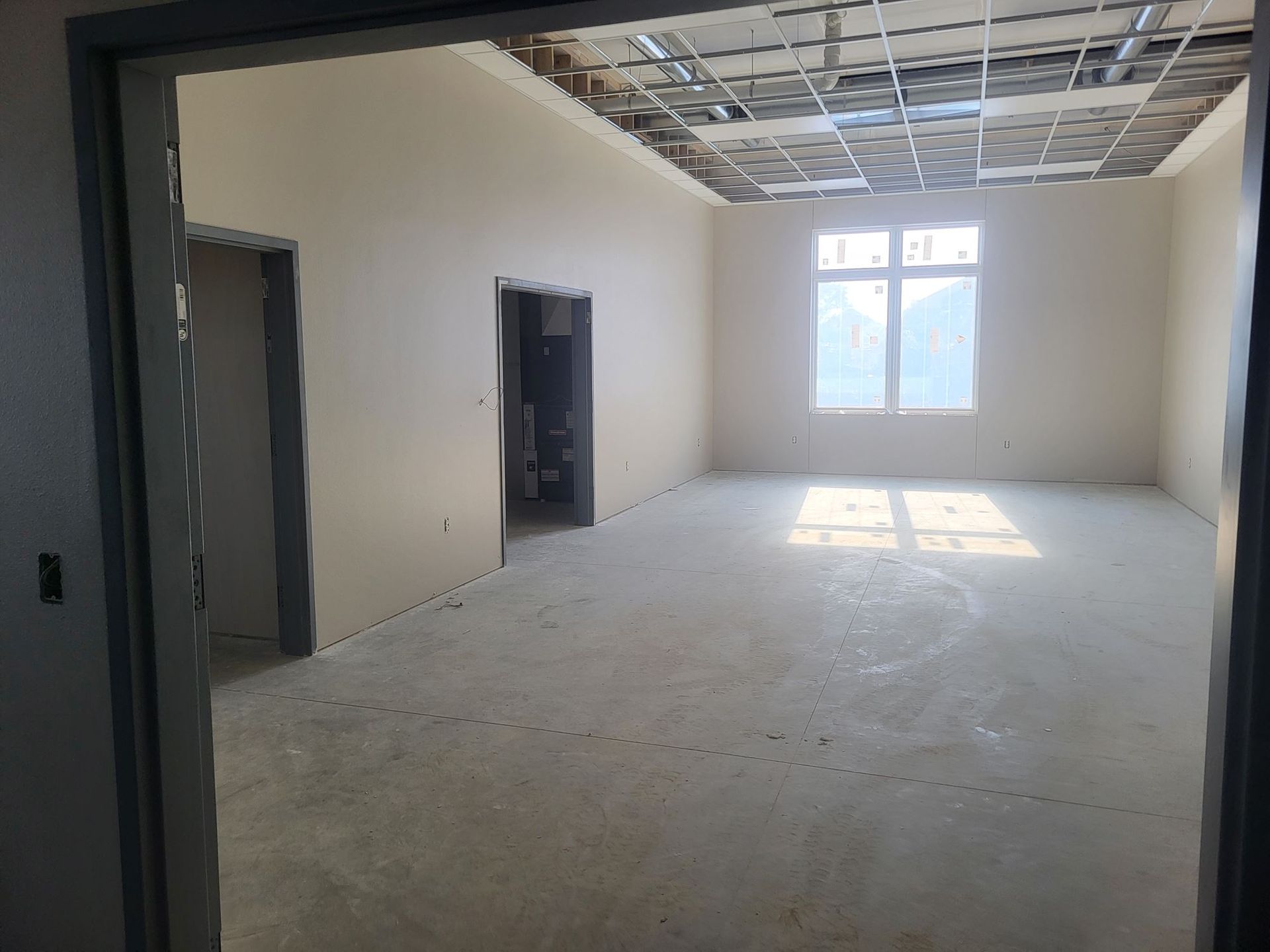 Empty unfinished room with bare walls, exposed ceiling grid, and sunlight through a window.