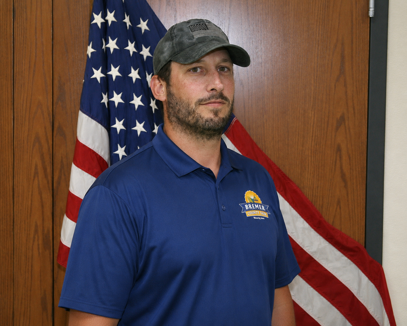 Man in red shirt and cap standing before wood paneling with an American flag behind him