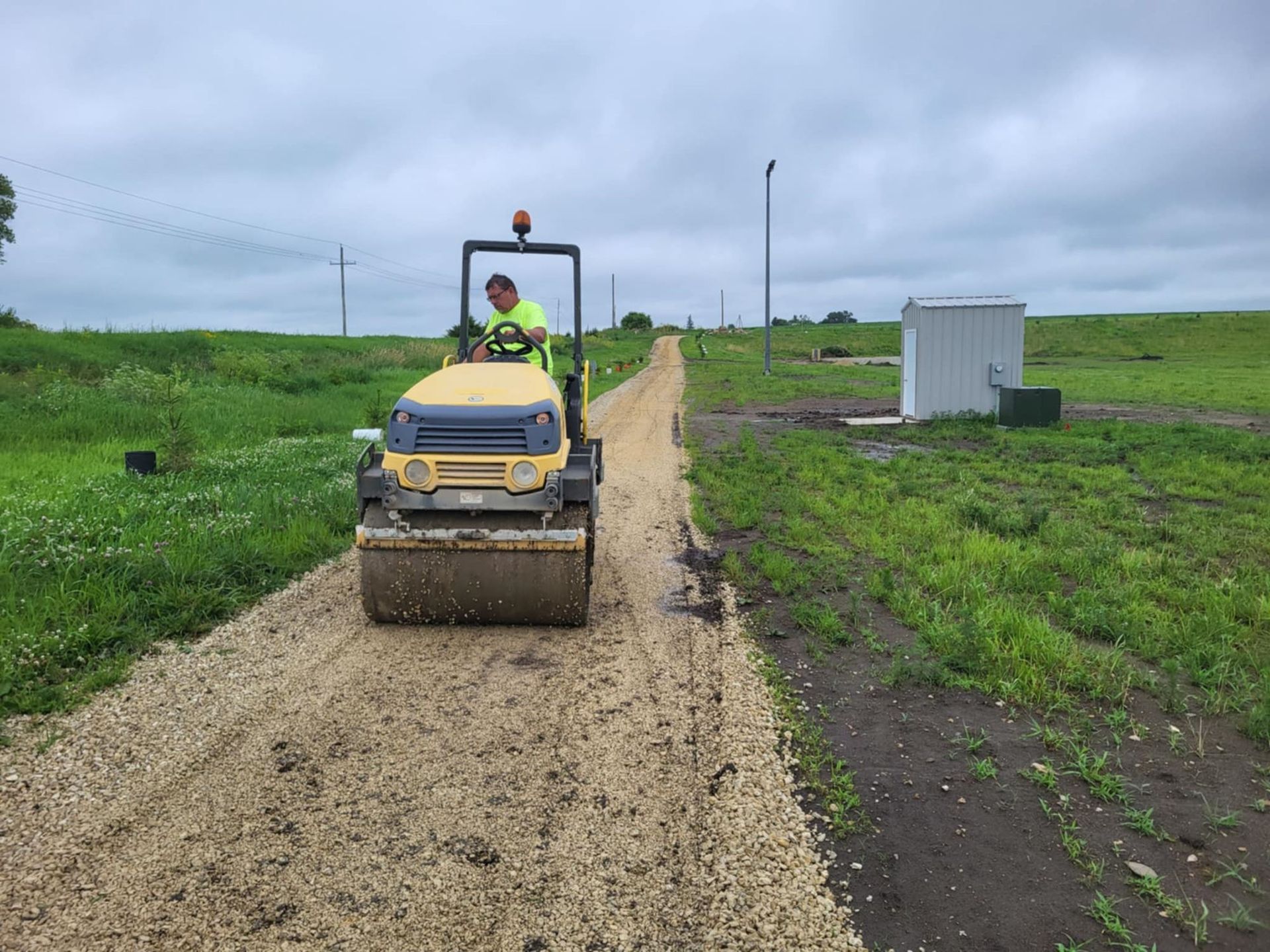 Small construction roller grading a dirt path across a green field under cloudy skies.