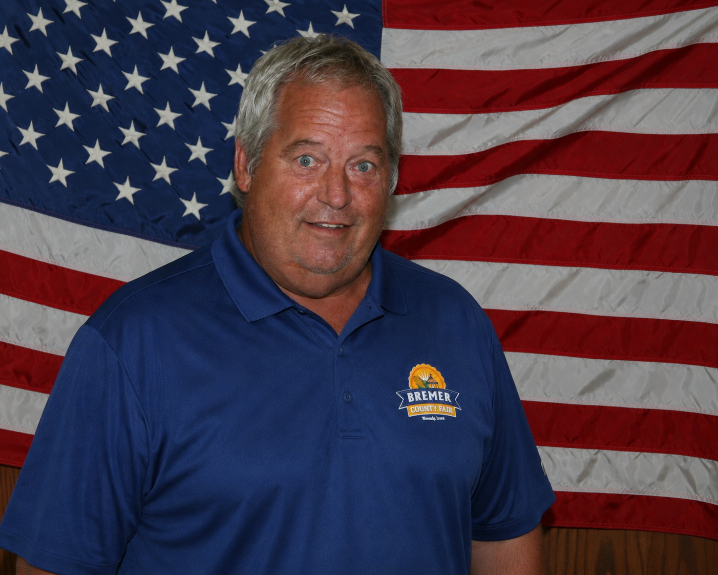 Man in red shirt smiling in front of a wood-paneled wall with an American flag.