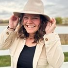 Smiling person in a beige hat and jacket, holding the brim outdoors by a wooden fence