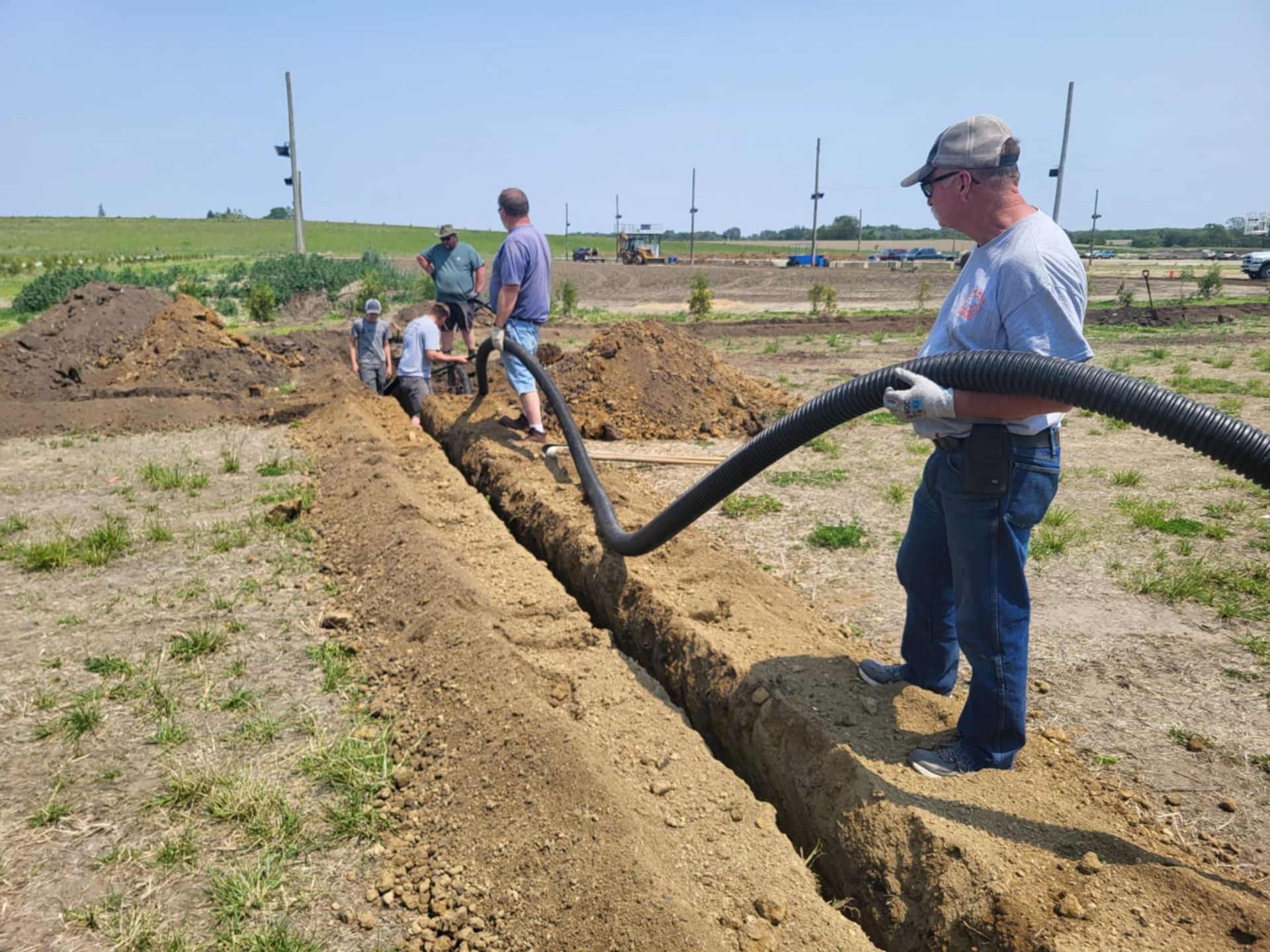 Workers lay underground pipe in a trench beside a dirt road in a rural field.