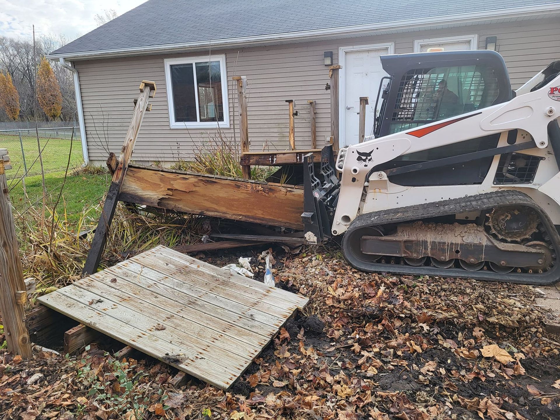 Compact tracked loader pushing debris near a house, with fallen panels and leaf-covered ground.