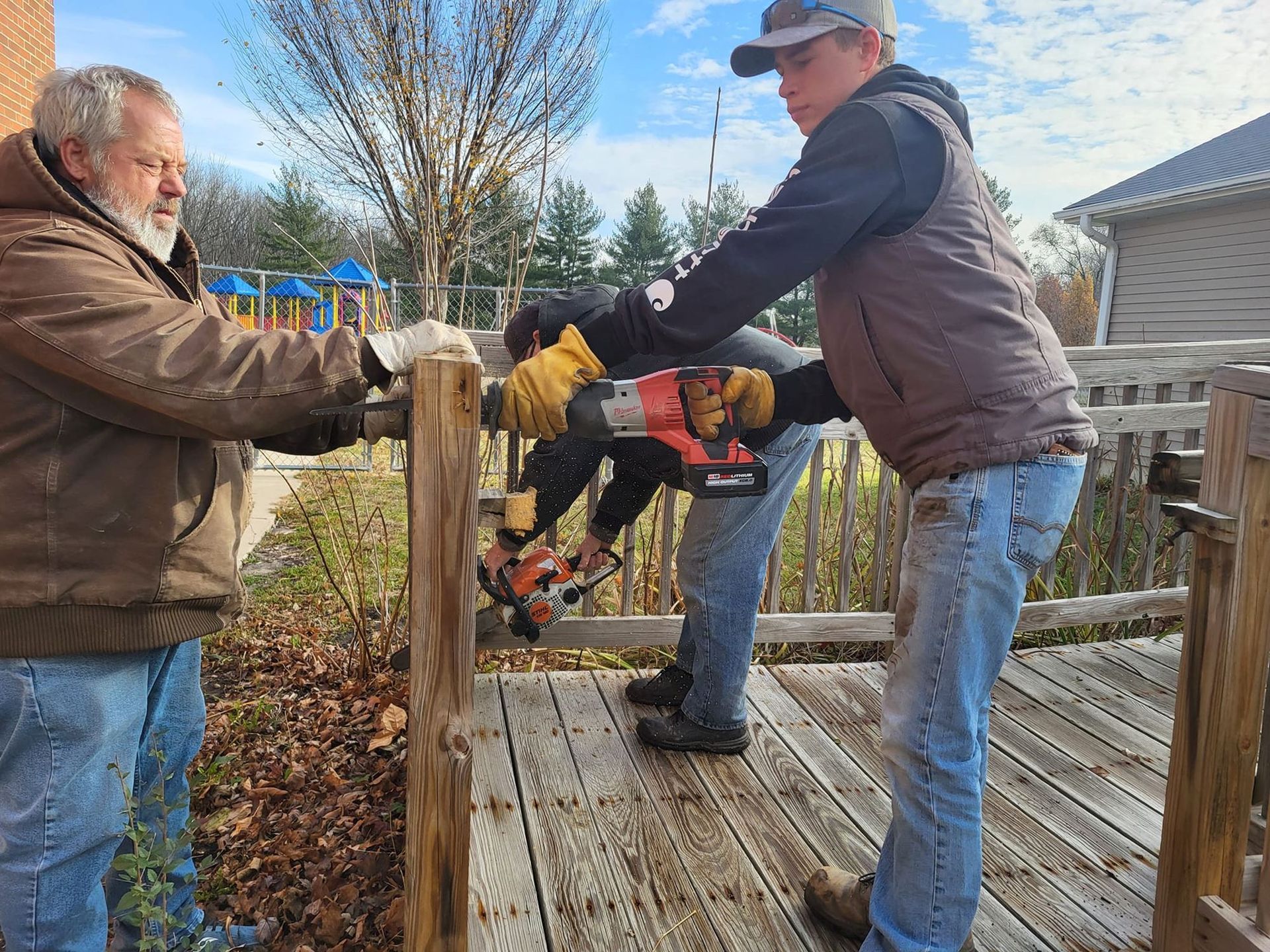 Two people using a chainsaw to cut a wooden post on an outdoor deck.