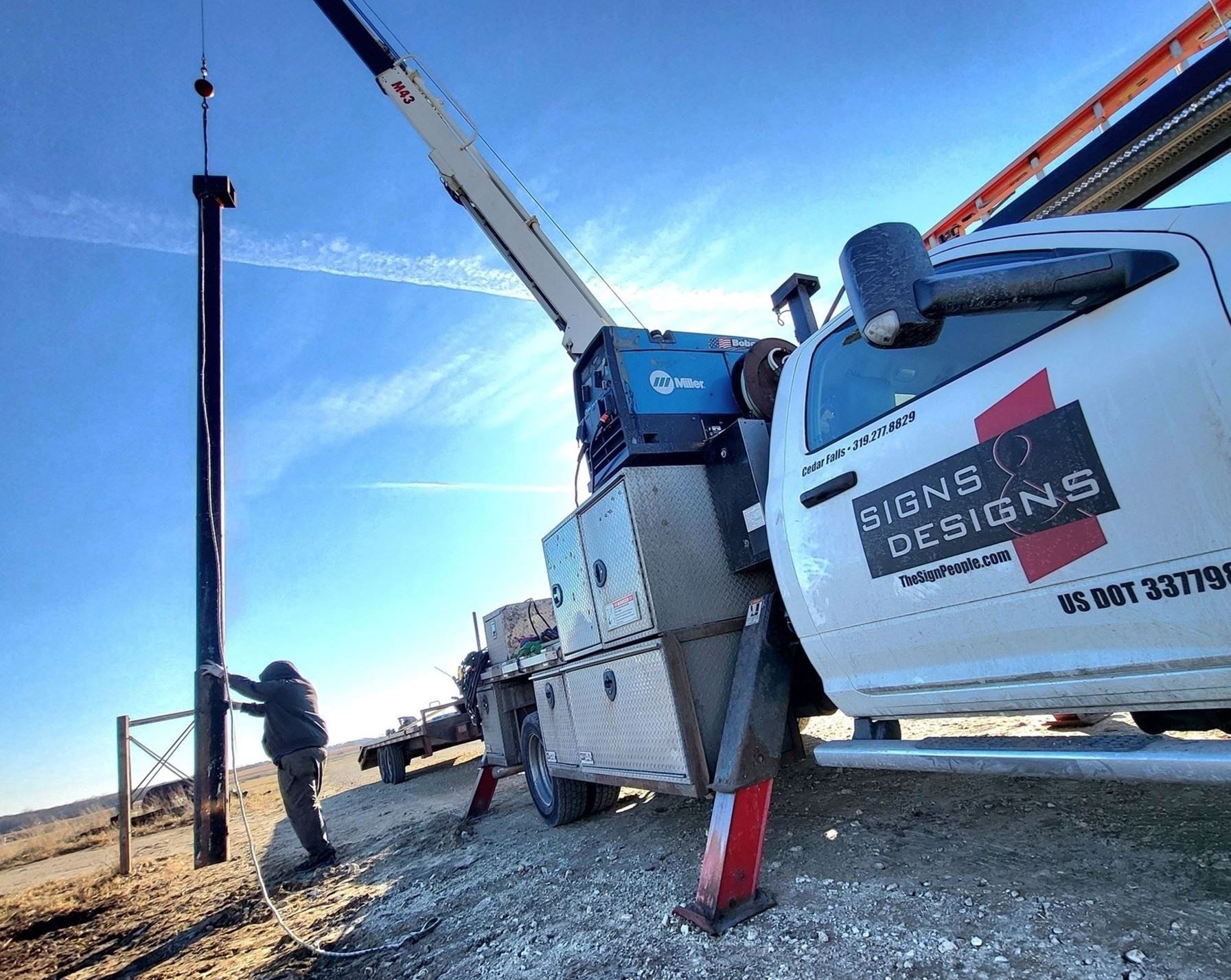 Crane truck lifting a utility pole while a worker guides it on a gravel roadside under a blue sky