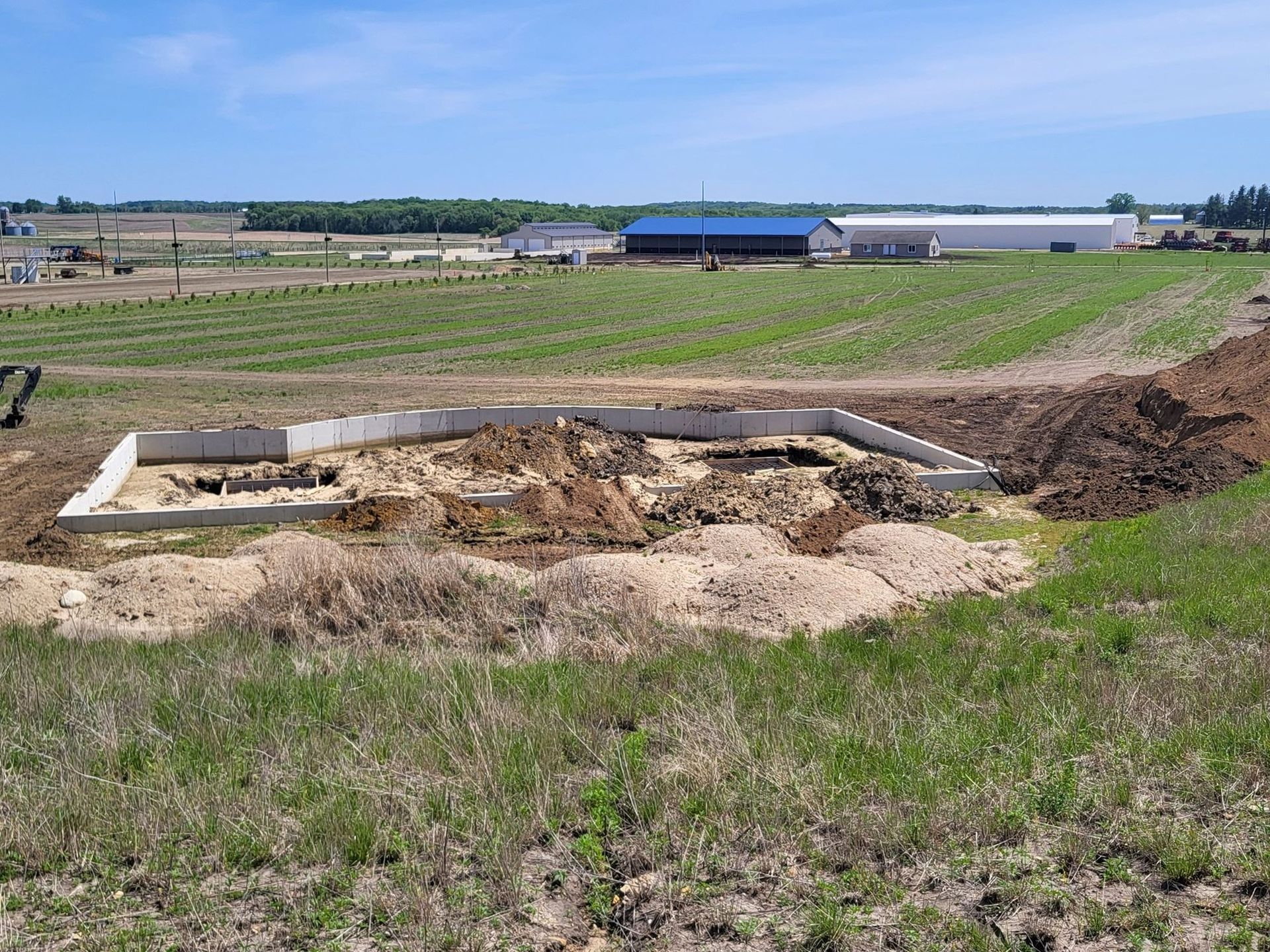 Excavated foundation pit in a grassy field, with dirt piles and buildings in the background.