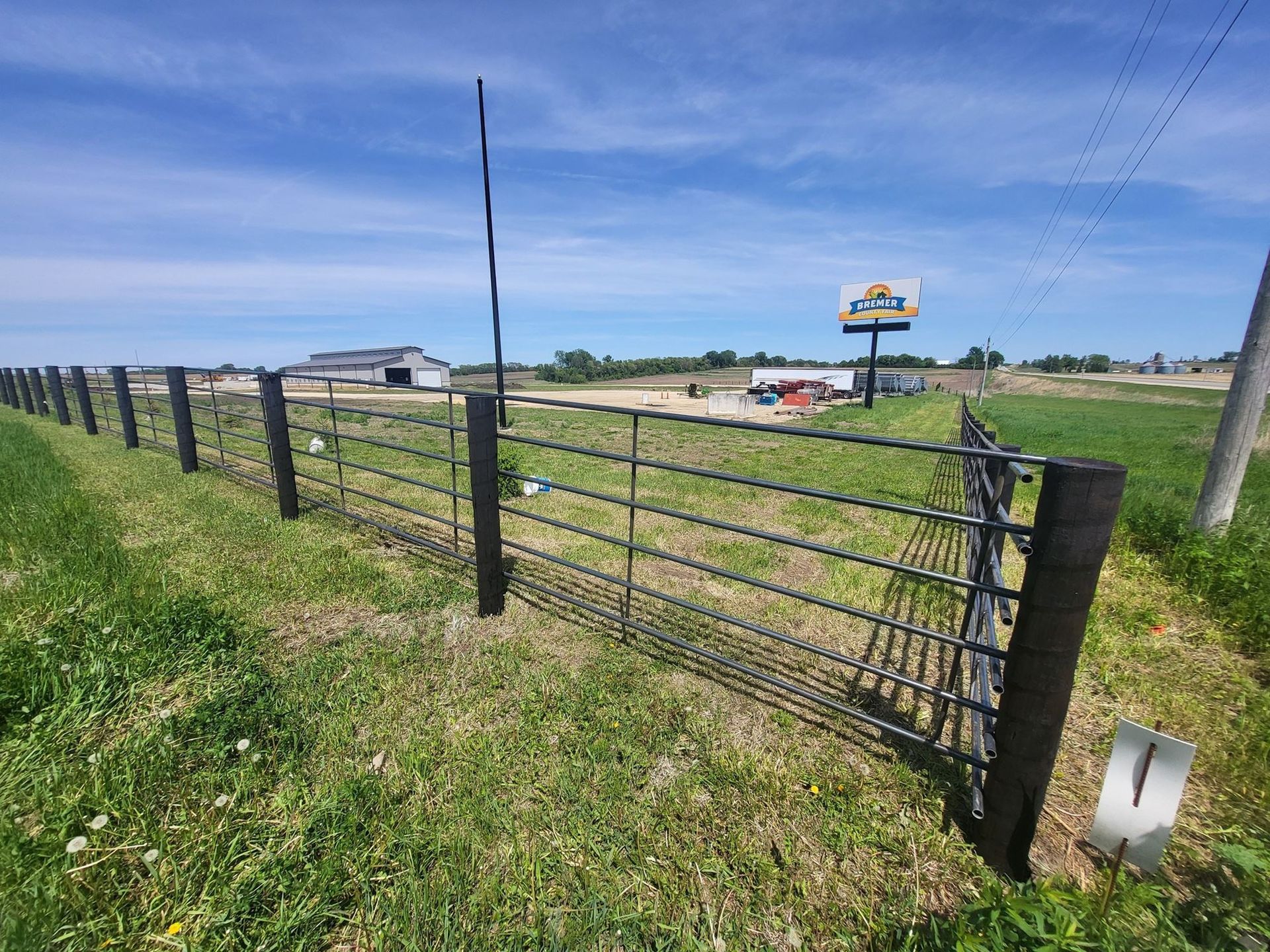 Rural roadside fence gate beside a grassy field under a blue sky, with buildings and a sign in the distance.