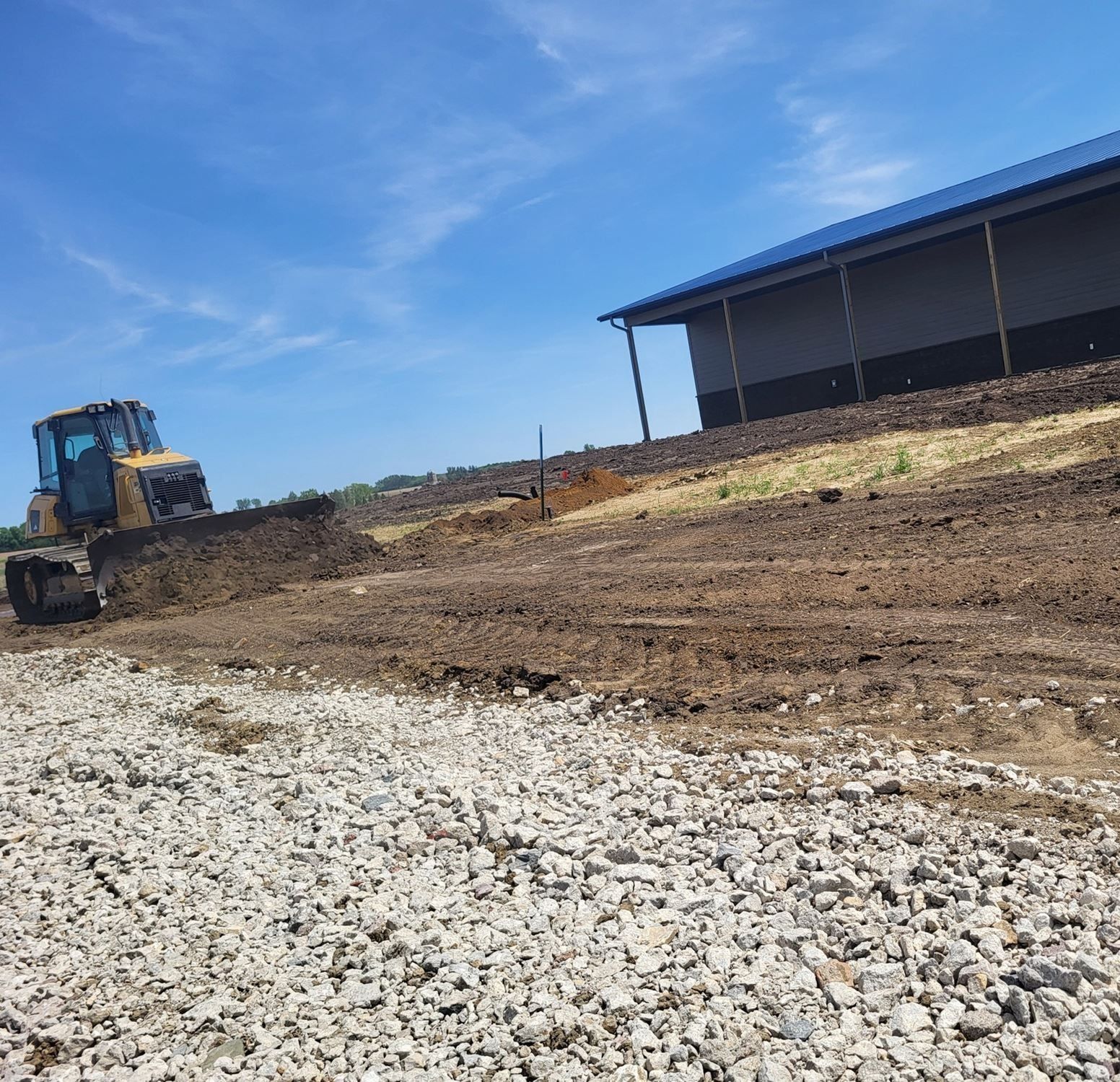 Bulldozer grading dirt near a large blue-roofed metal building on a sunny day