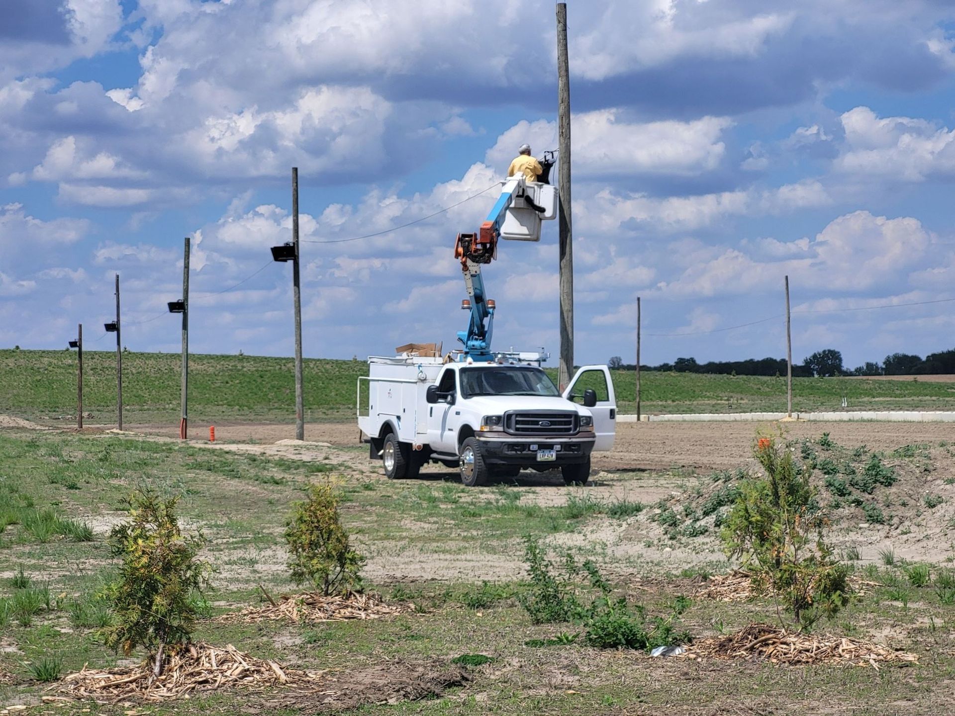 Utility truck with bucket lift and worker trimming poles in a rural field under a cloudy sky