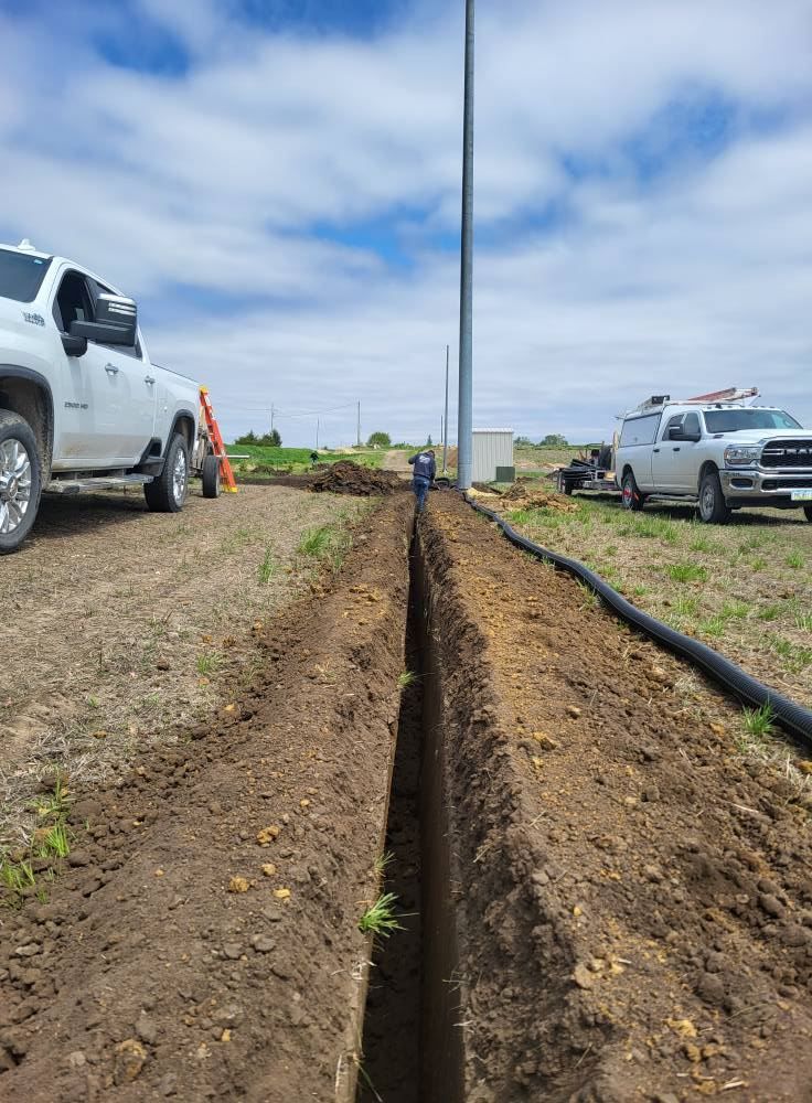 Trench dug through dirt between parked trucks under a blue sky, with a pipe alongside it.