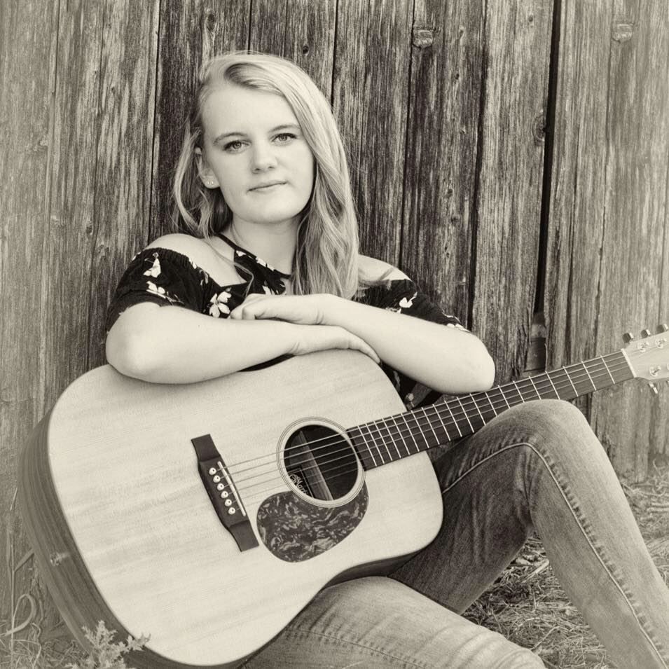 Woman sitting with an acoustic guitar against a wooden wall, black-and-white portrait