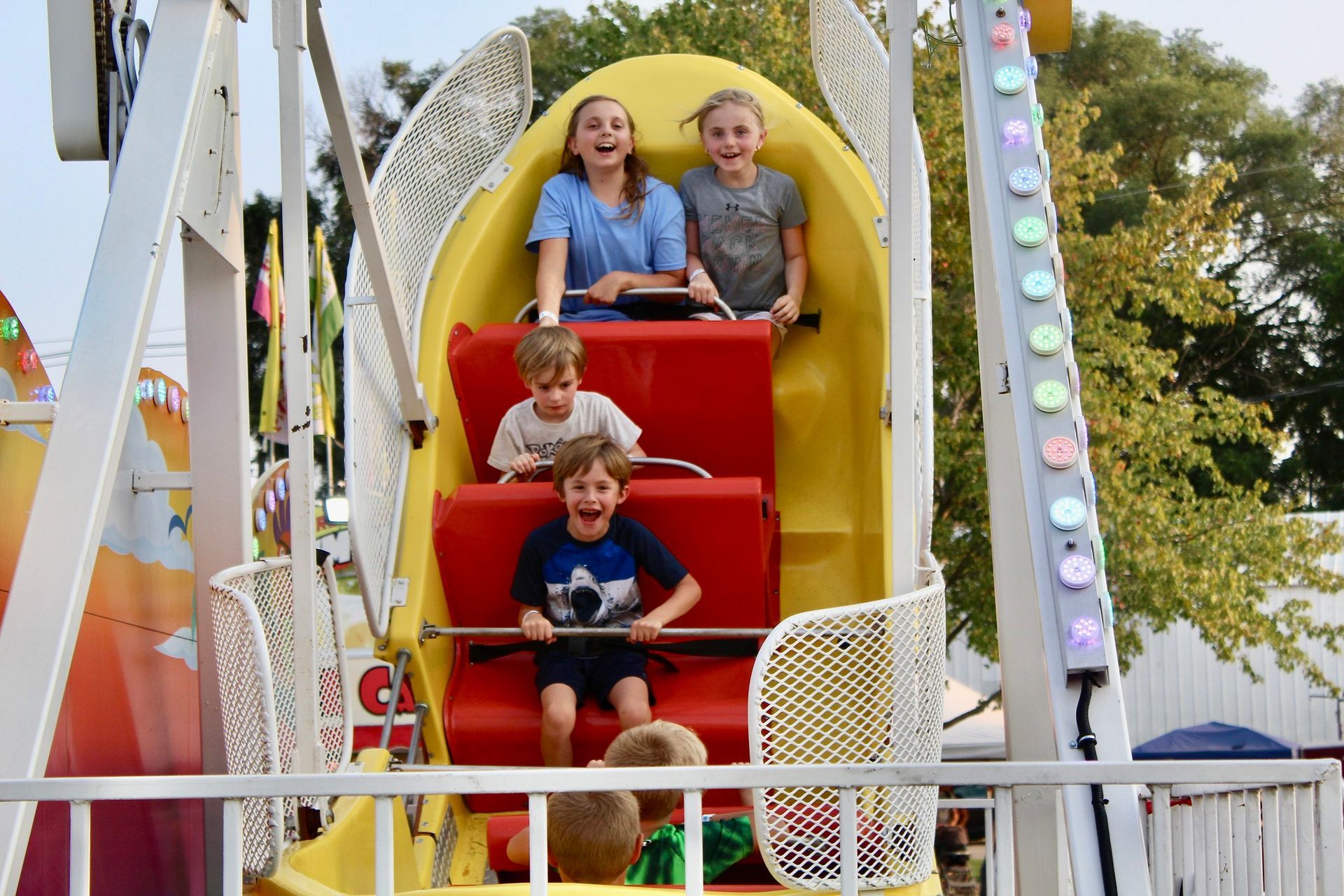 Two children and two adults riding a yellow Ferris wheel seat, smiling at a fair.