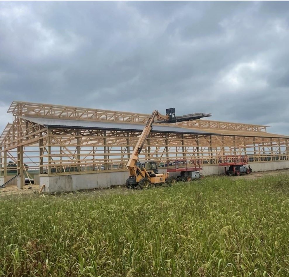 Construction equipment lifting a wooden building frame in a grassy field under cloudy skies