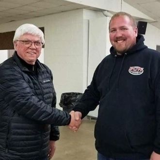 Two men in jackets shake hands indoors, smiling at the camera.