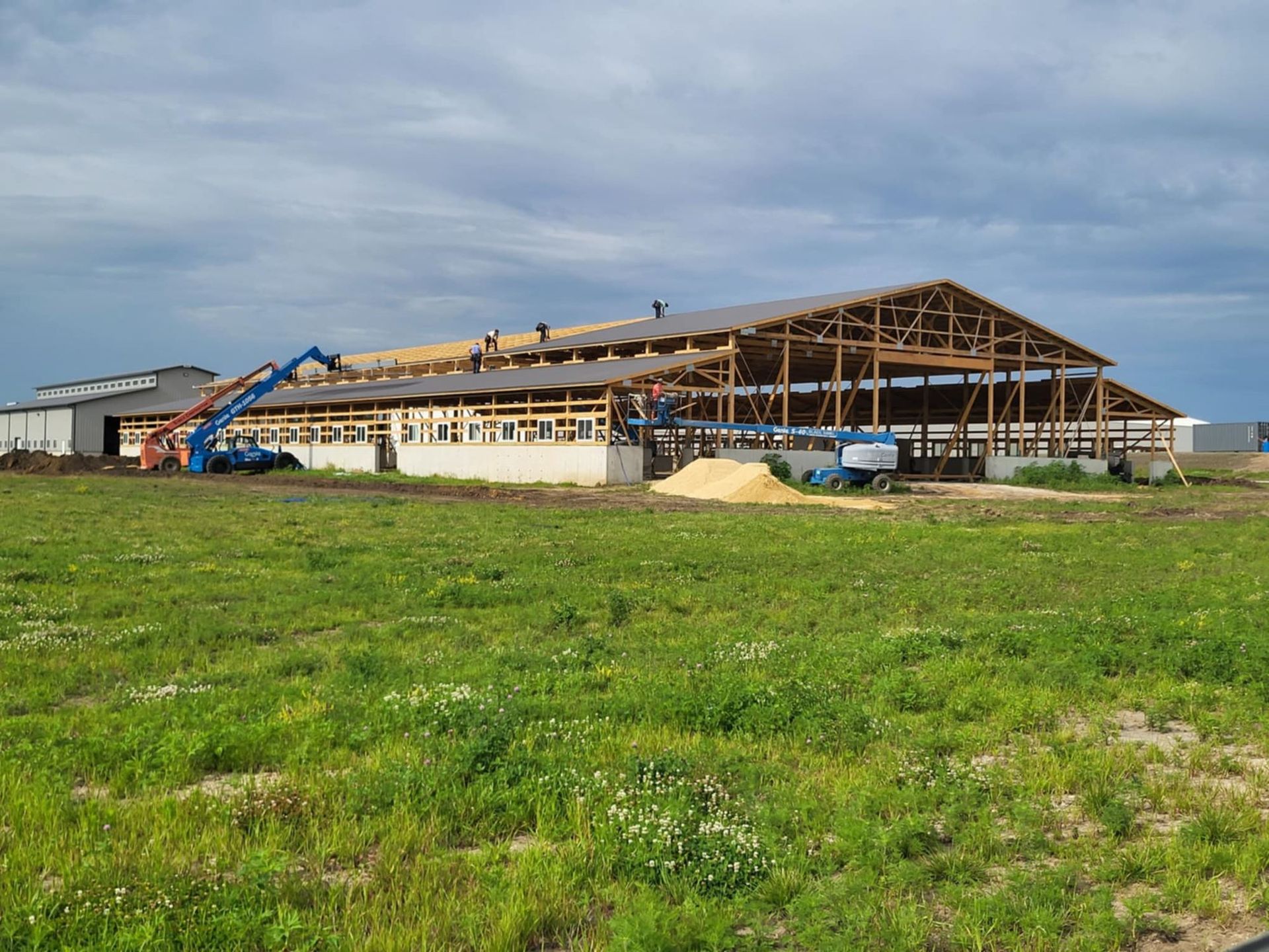 Large wooden building under construction in a grassy field, with a blue crane and open roof framing.