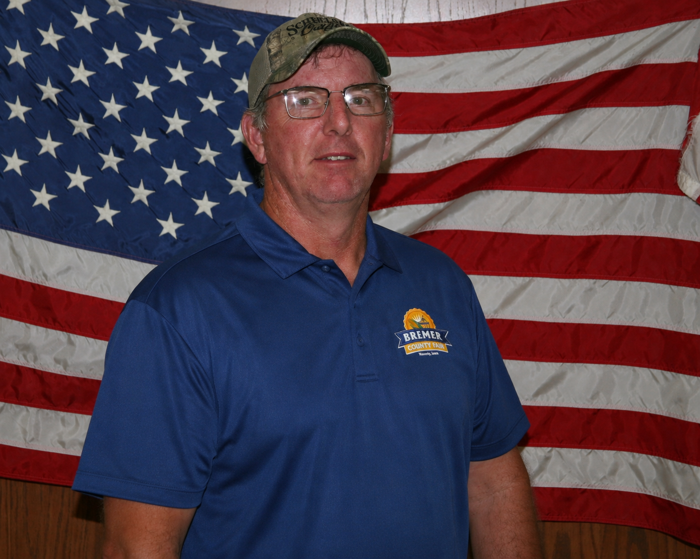 Man in blue sweatshirt and cap standing before a U.S. flag and wood-paneled wall