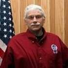 Man in red shirt standing before a wood-paneled wall with an American flag behind him