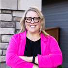 Smiling person with glasses and blonde hair, wearing a bright pink cardigan, standing outdoors by a house wall.