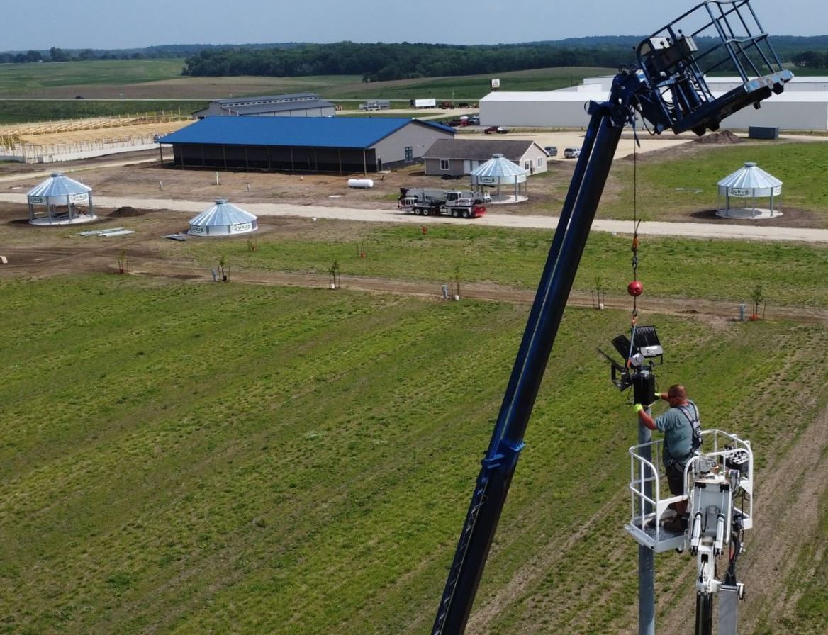Workers on a tall boom lift inspecting equipment beside a grassy field and blue-roofed buildings