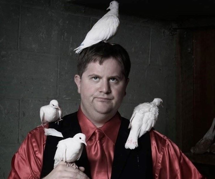 Man in red shirt and tie with several white birds perched on his head and shoulders against a dark background