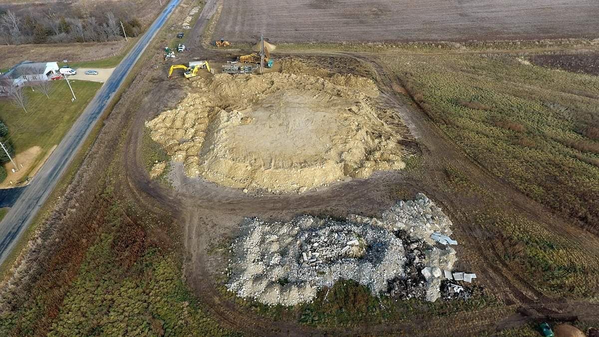Aerial view of two large dirt mounds beside a rural road and open fields.