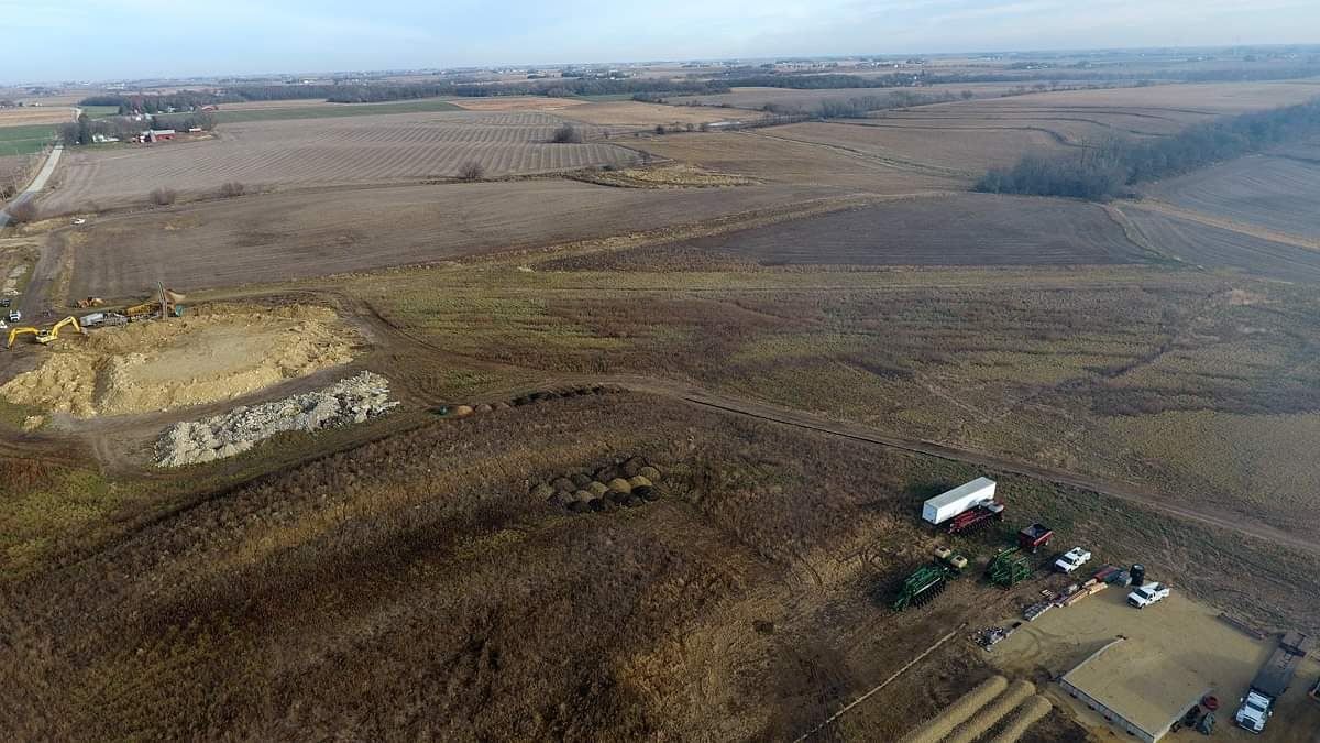 Aerial view of a muddy construction site beside open fields, with heavy equipment and dirt piles.