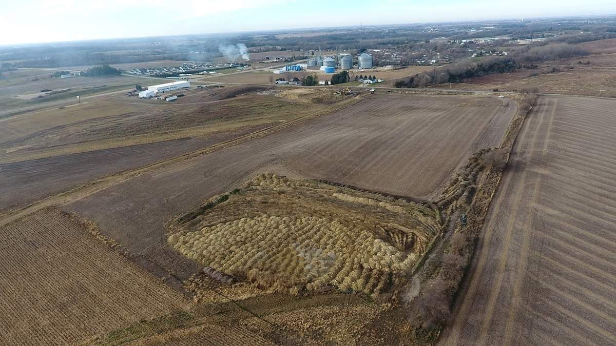 Aerial view of rural farmland with brown fields, a patch of grass, and distant village buildings
