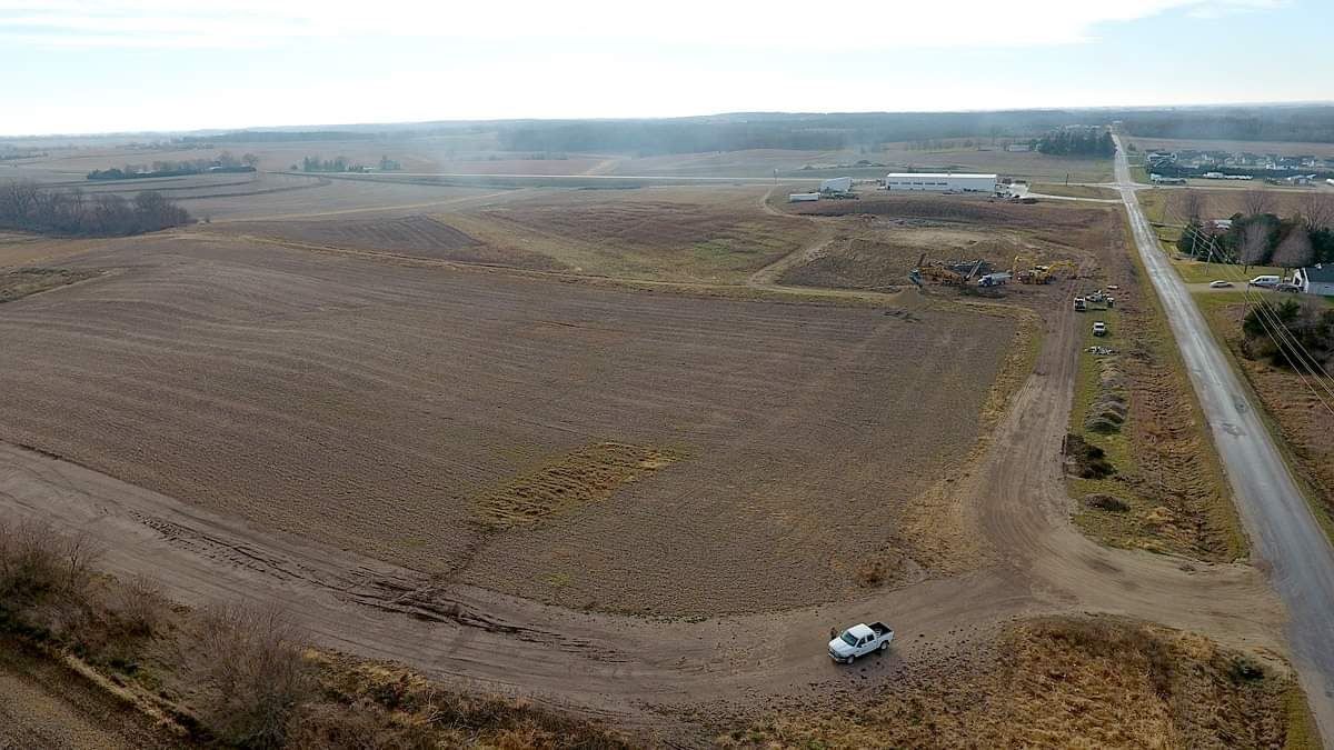 Aerial view of a large barren field beside a road with a parked white vehicle and distant buildings.