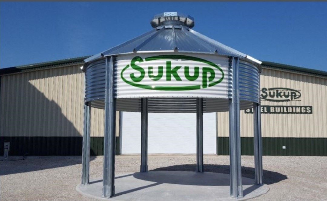 Sukup grain bin display with green logo outside a metal building under a clear sky