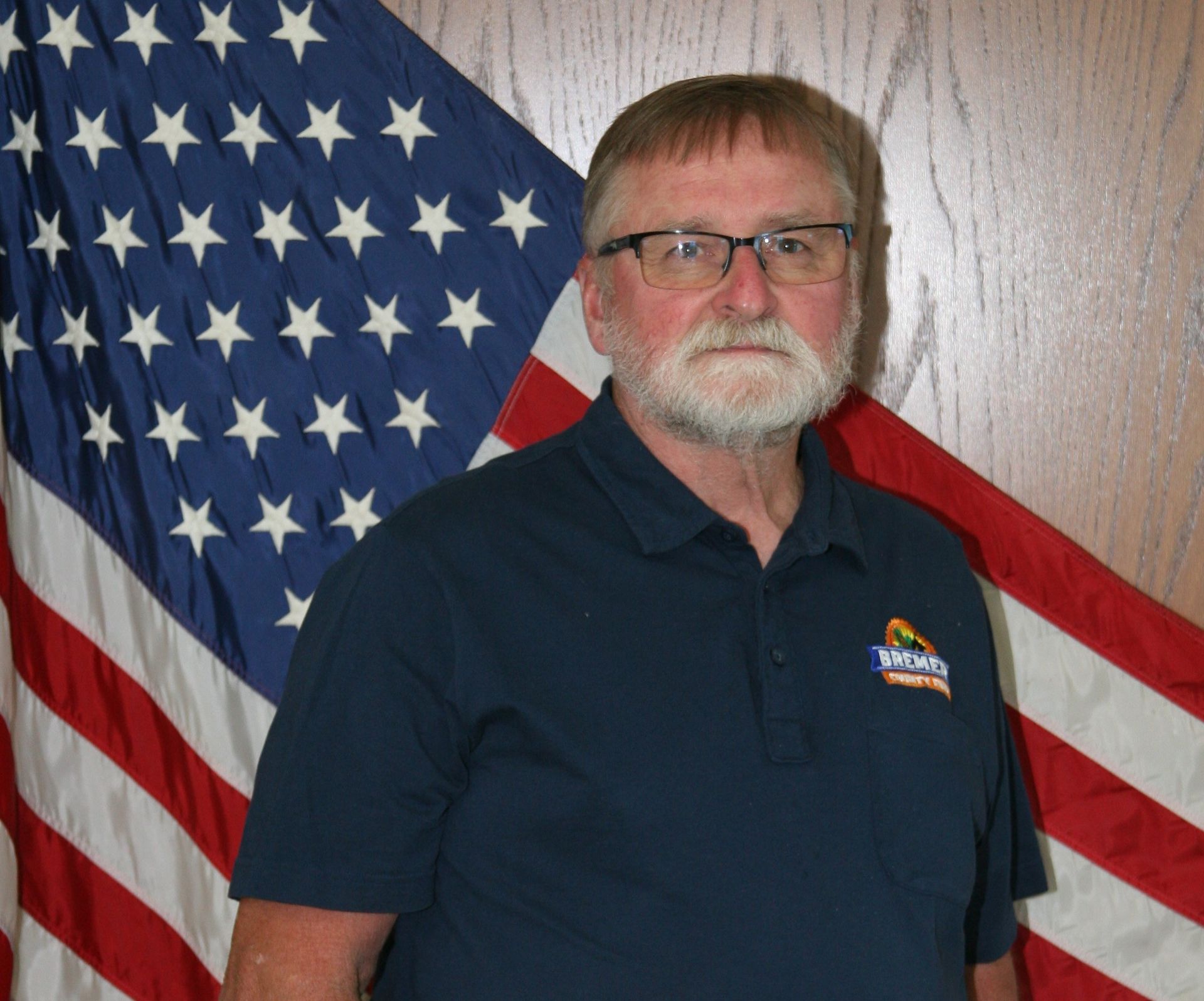 Man in a red shirt standing before a wood-paneled wall and U.S. flag