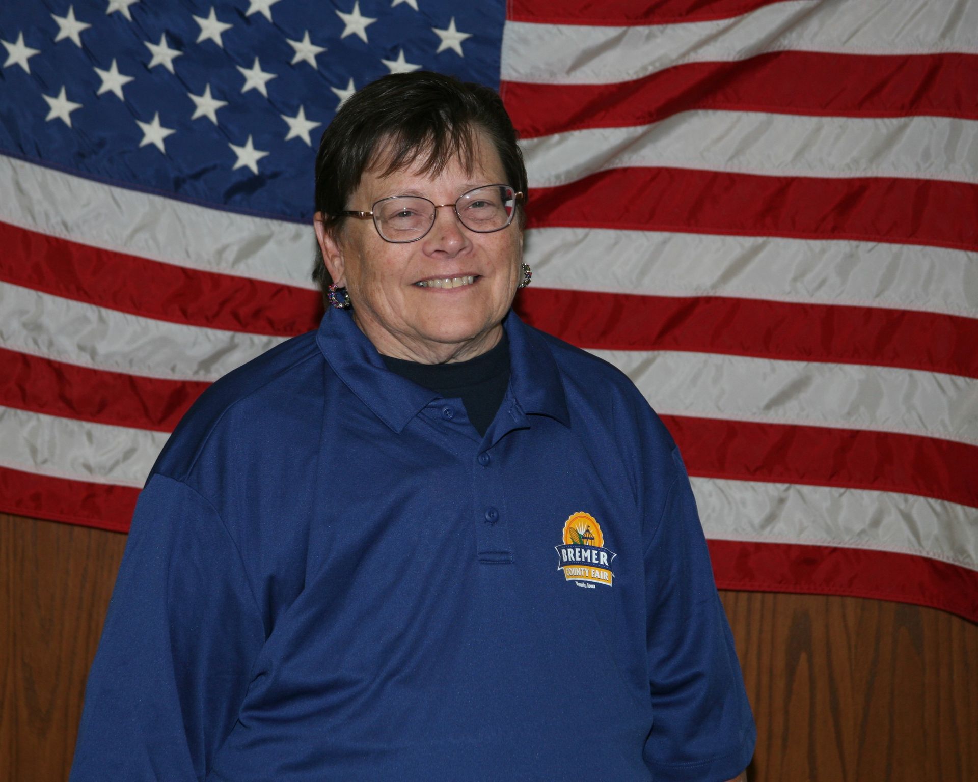 Smiling person in a red shirt standing before an American flag and wood paneling