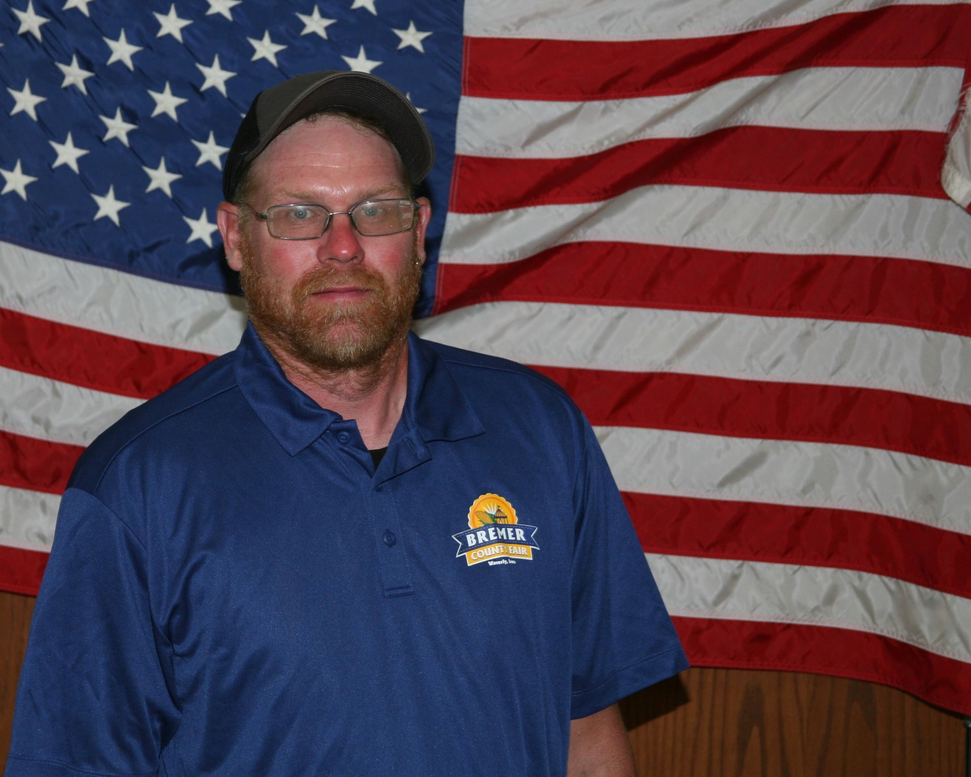 Man in glasses and green cap wearing a dark shirt, standing indoors against a plain wall.