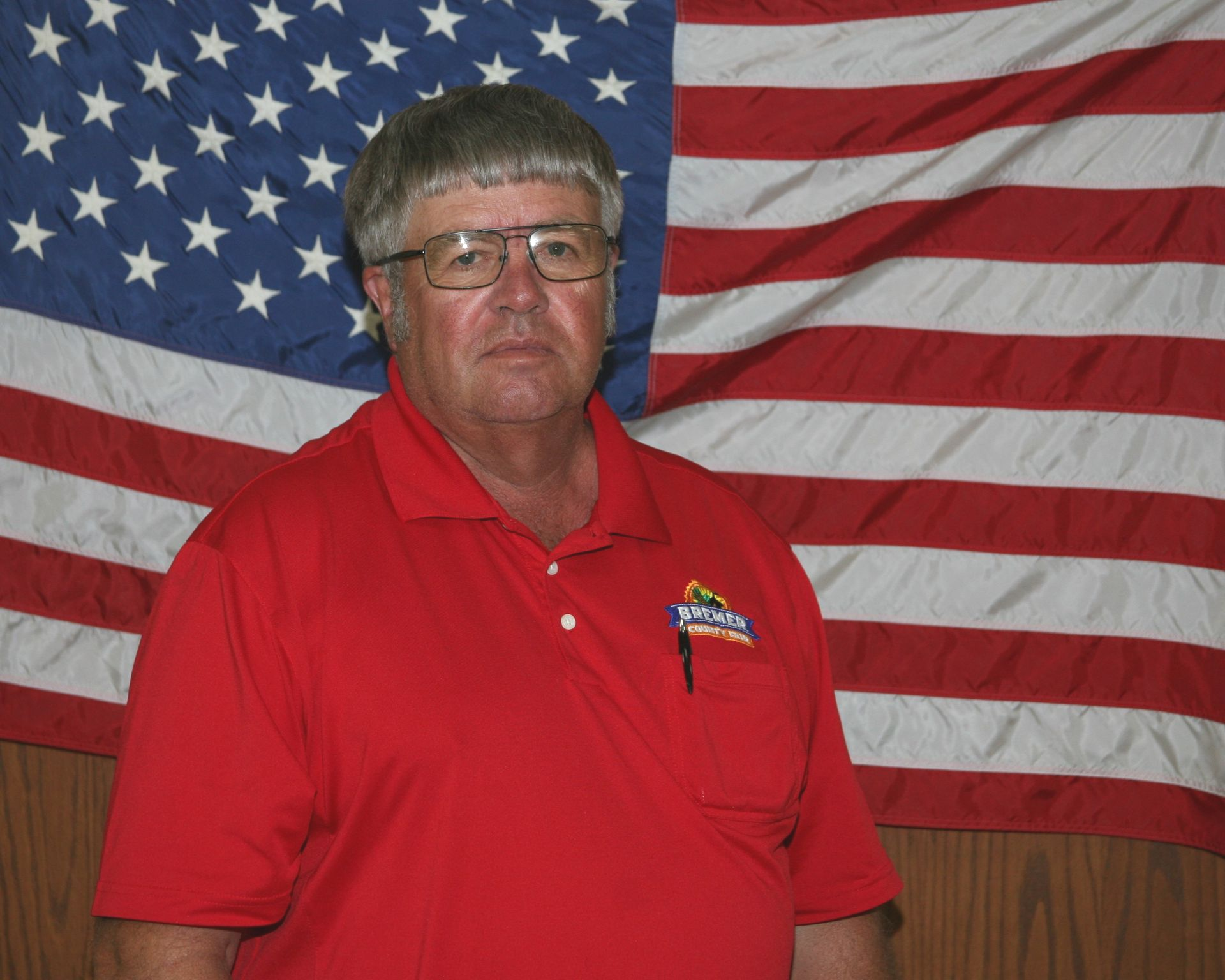 Man in a red polo shirt standing against a wood-paneled wall, wearing a small emblem badge