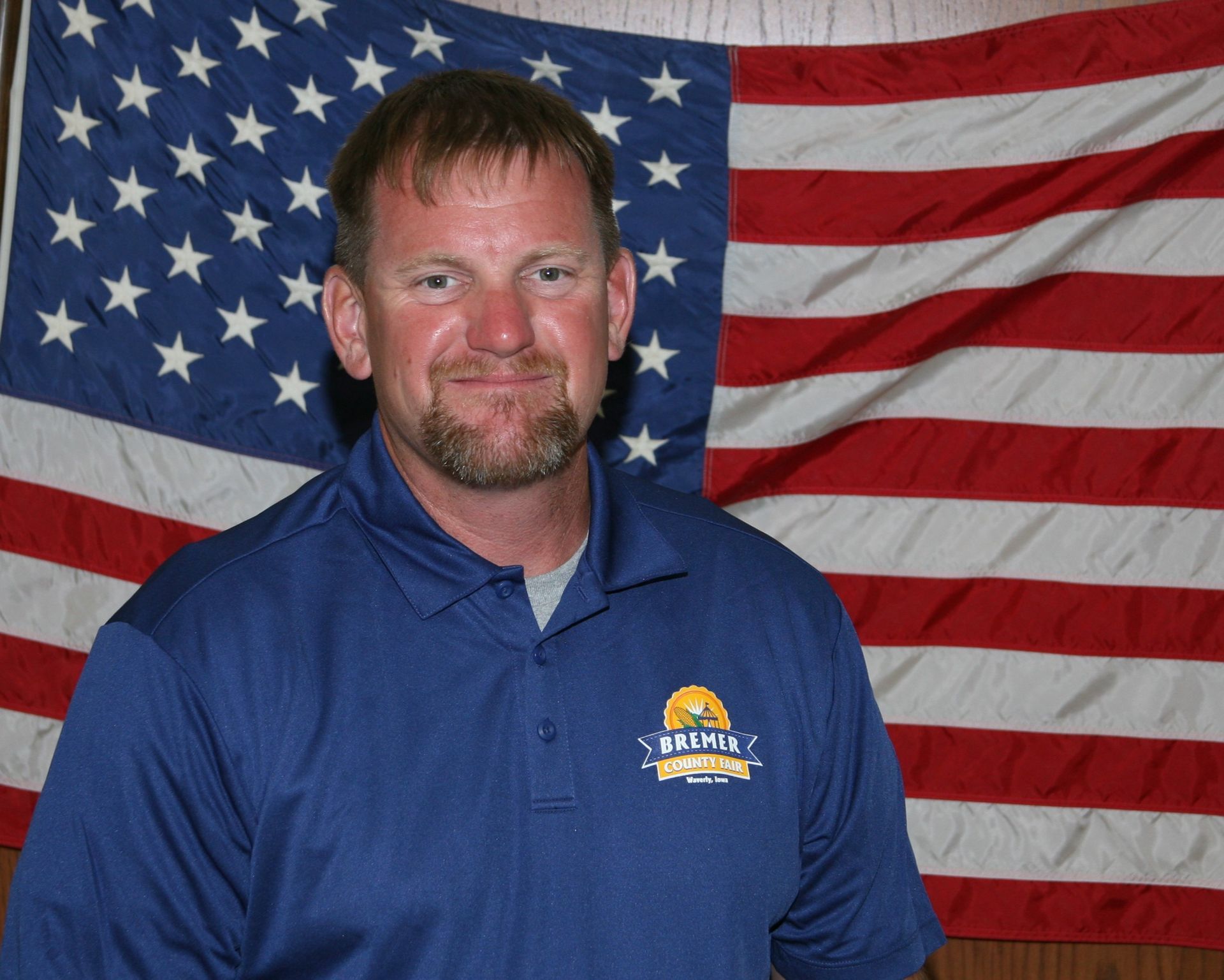 Man standing indoors before wood paneling and a U.S. flag, wearing a navy polo shirt.