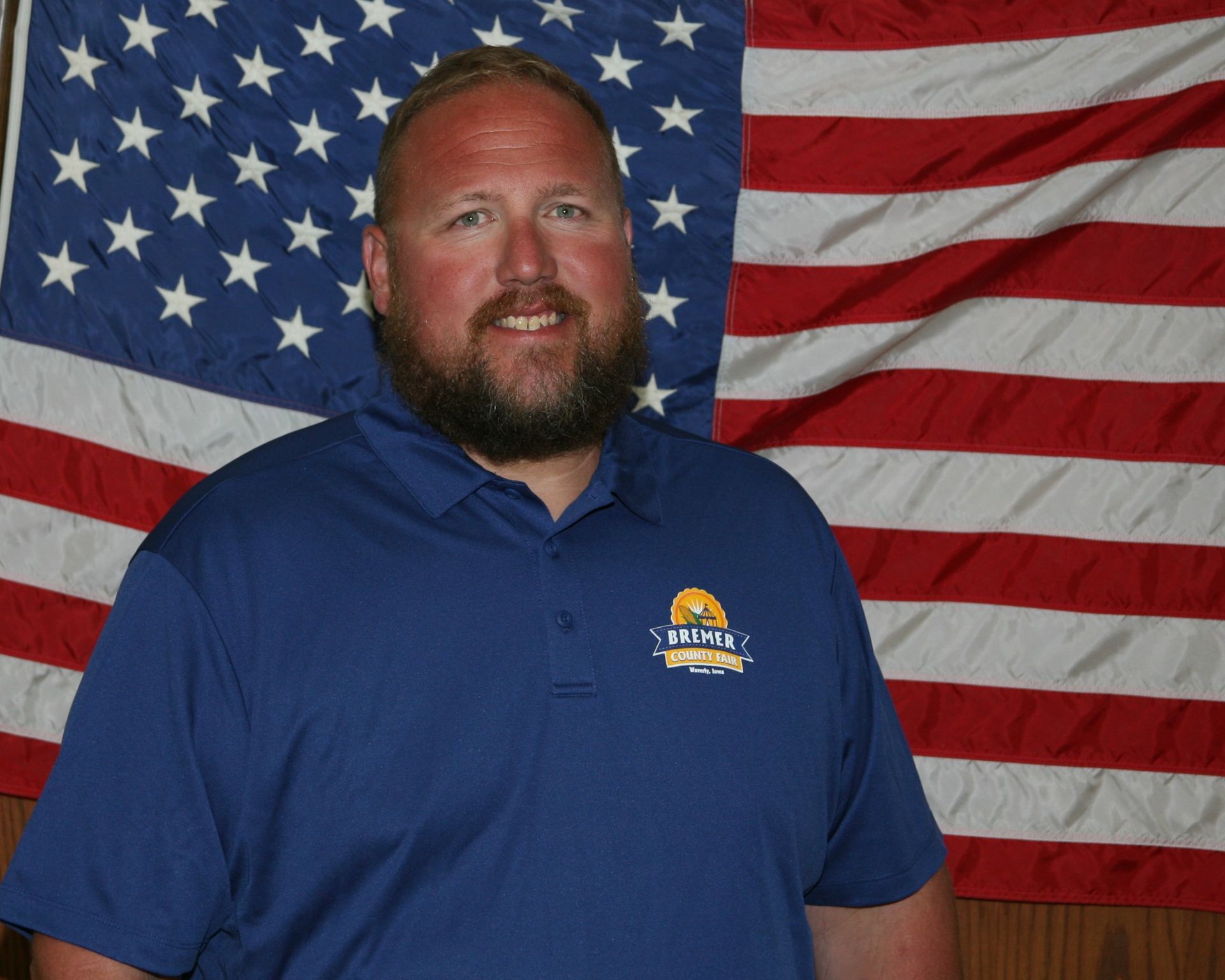 Smiling man in a red shirt standing before an American flag and wood-paneled wall