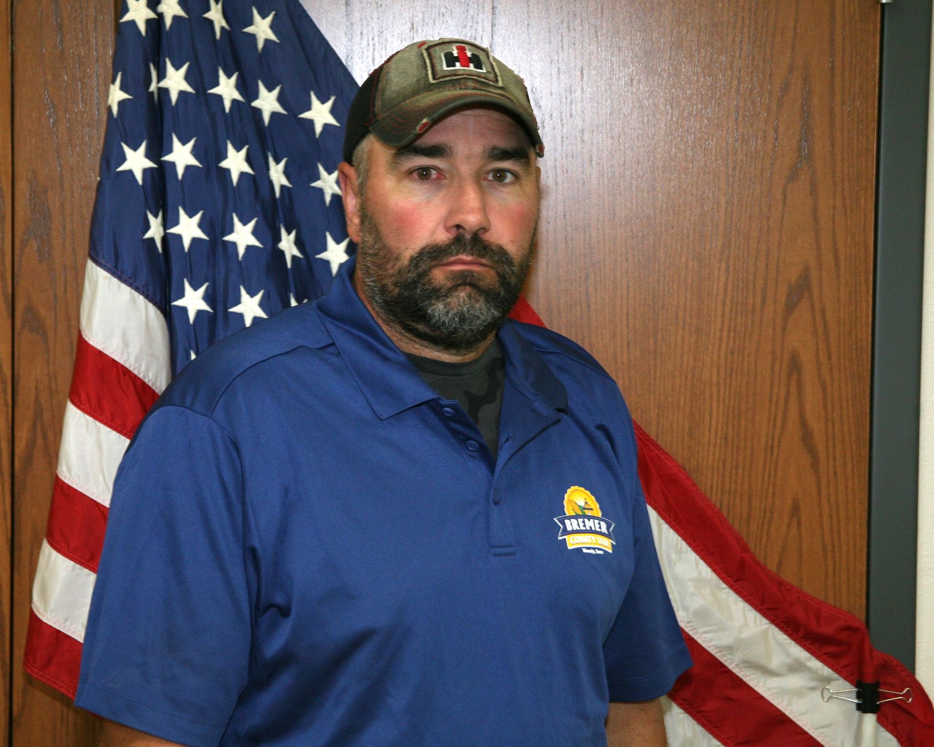 Man in a red button-up shirt and green cap stands indoors beside a U.S. flag.