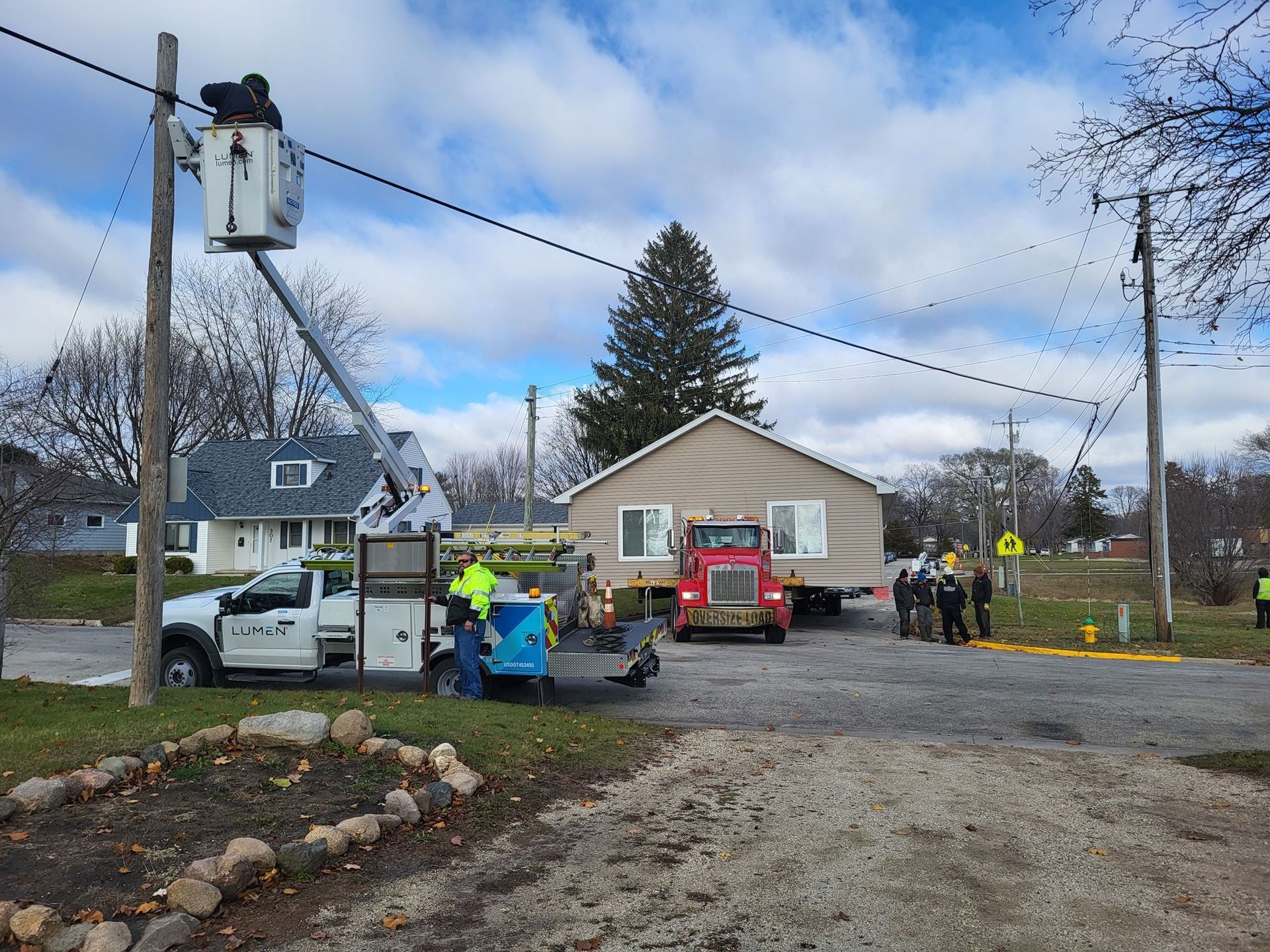 Utility workers repairing a downed power line beside a house, with trucks and cones on a gravel street