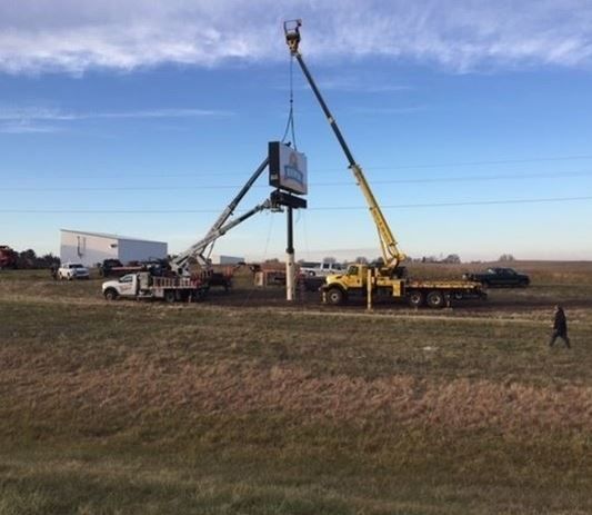 Construction cranes lifting a blue structure beside trucks in a grassy field under a clear sky