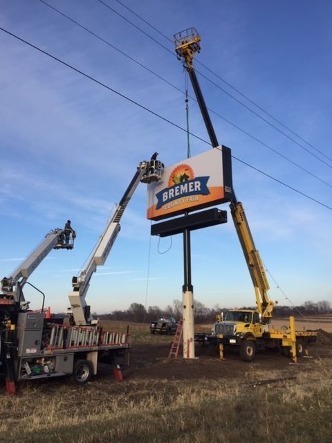 Crane crews lift a large roadside sign beside a highway under a blue sky