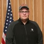 Person in a black jacket and cap standing in front of an American flag and wood-paneled wall