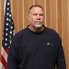 Man standing before wood paneling and an American flag, wearing a dark sweatshirt with a serious expression