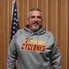 Man in gray Iowa State Cyclones hoodie standing indoors in front of wood paneling and an American flag