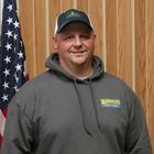 Man in a dark hoodie and cap standing indoors beside an American flag and wood-paneled wall
