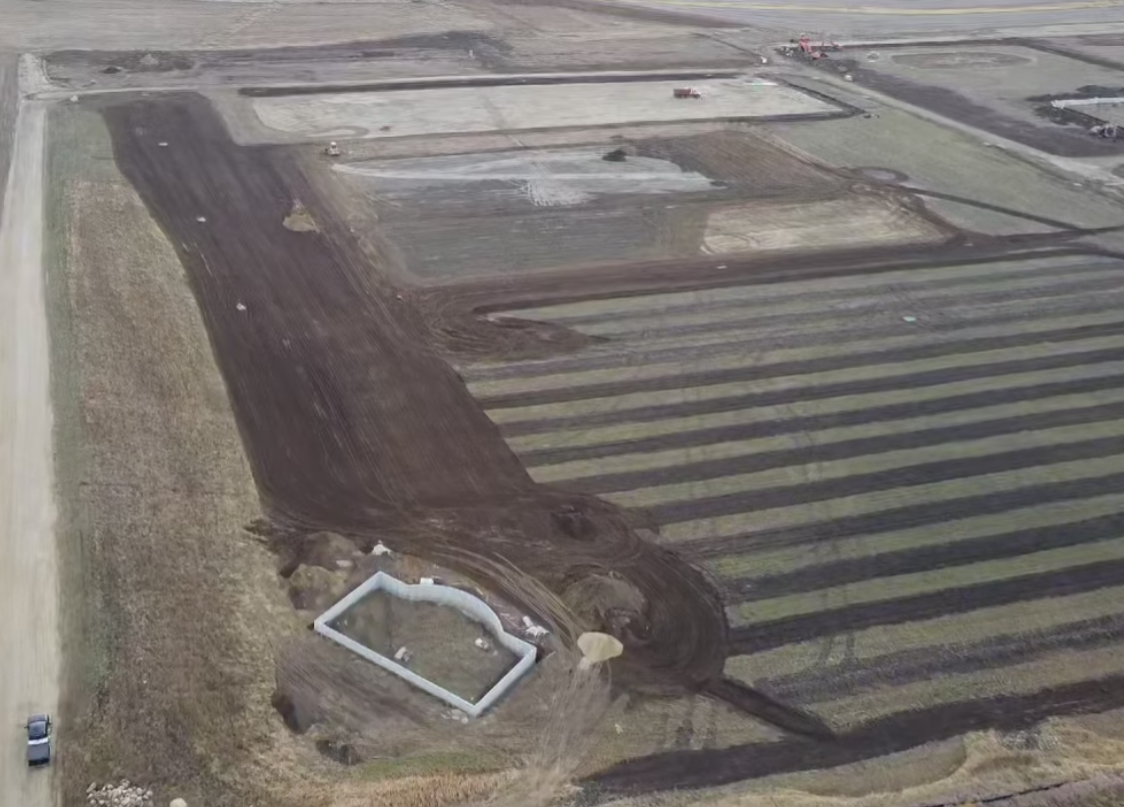 Aerial view of a construction site with dark soil strips, rectangular plots, and a small pond or basin.
