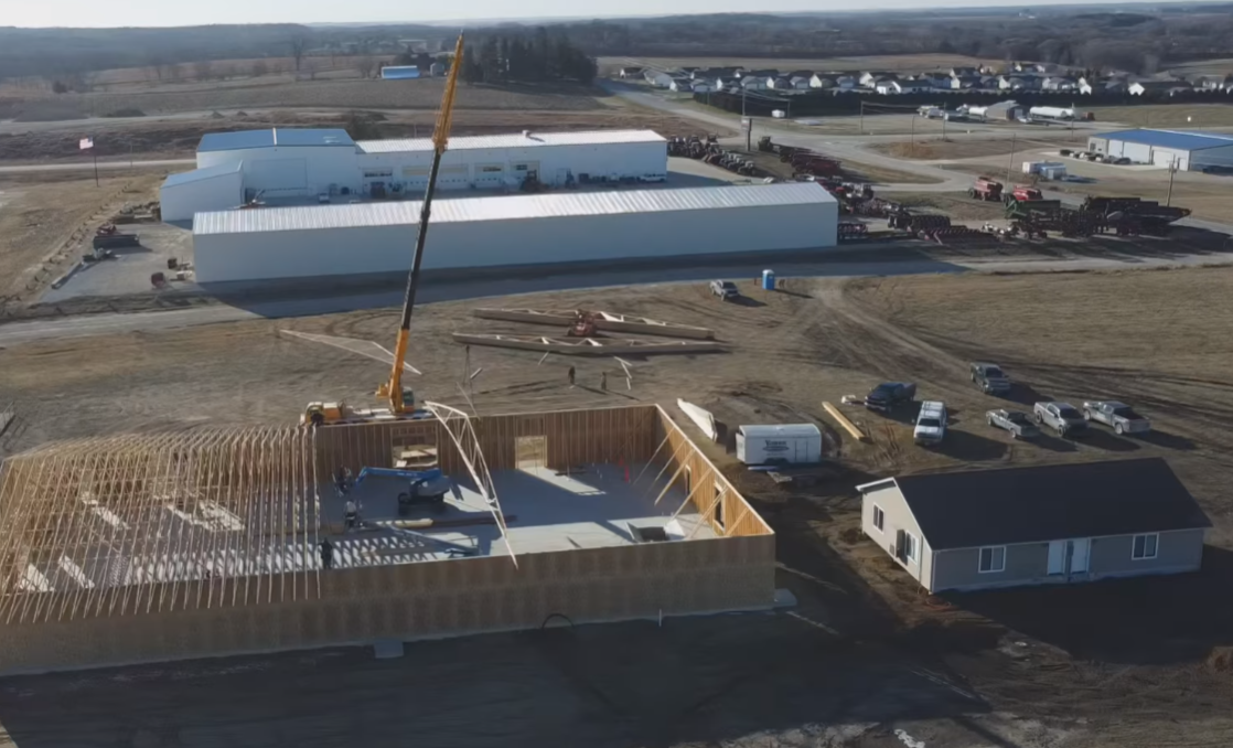 Aerial view of a construction site with a crane, partially built foundation, and large white industrial buildings nearby.