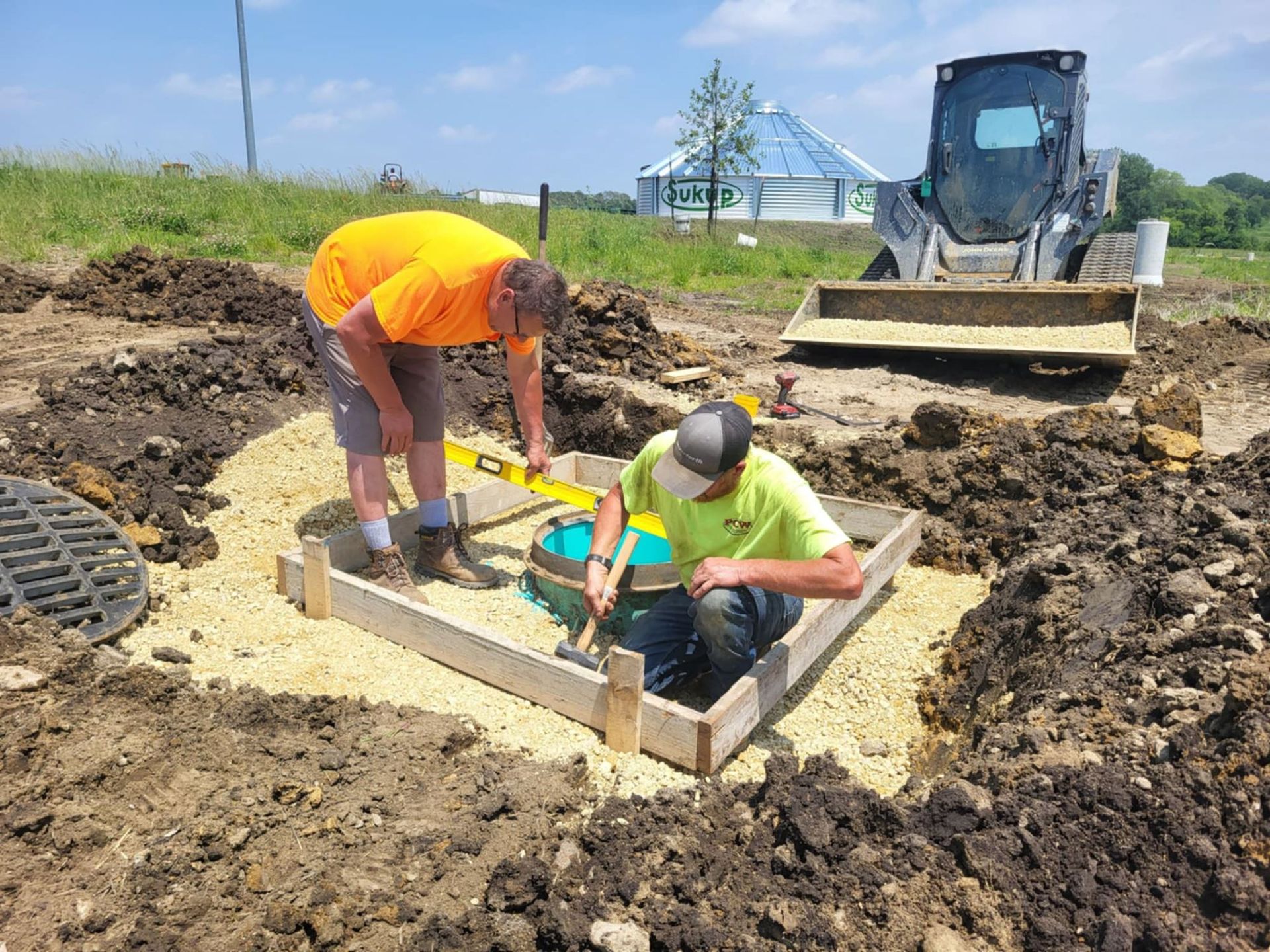 Two workers install a foundation frame in a muddy construction site beside a skid steer.