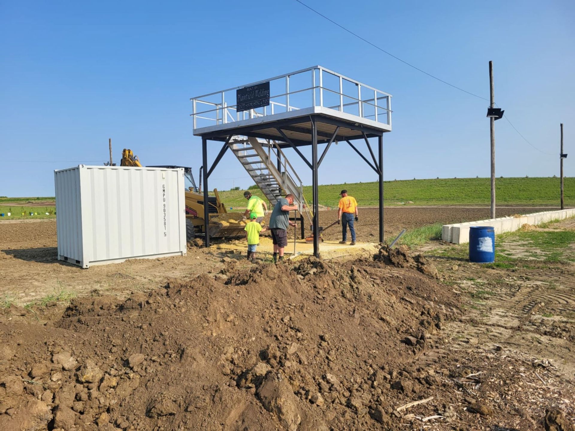 Workers at a dirt site beside a raised metal platform, with a white container and blue barrels nearby.