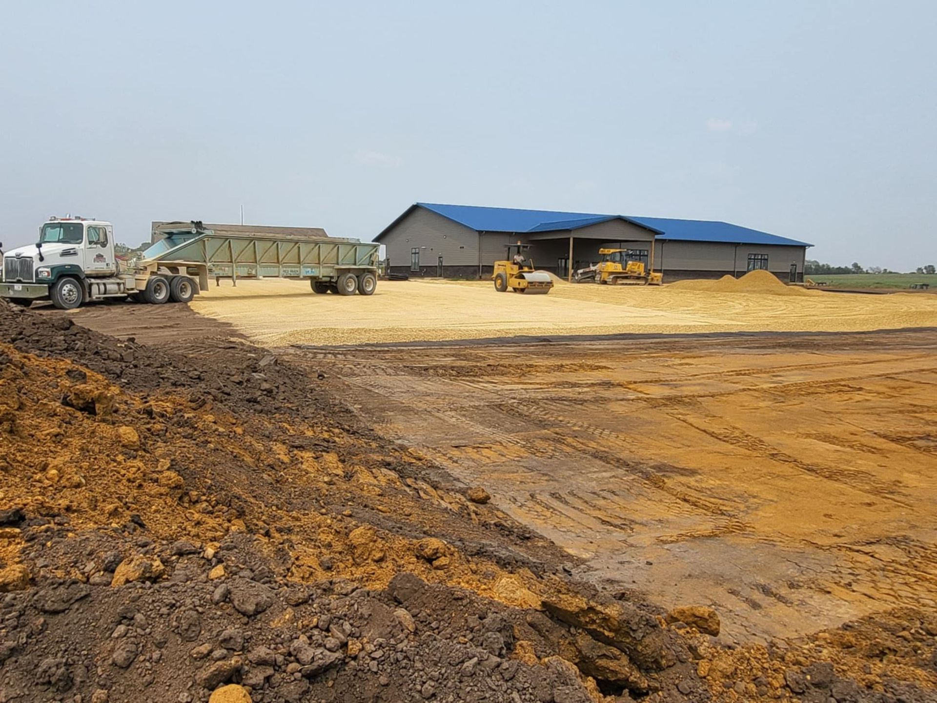 Construction site with earthmoving truck, trailer, and blue-roofed shed on a dirt lot