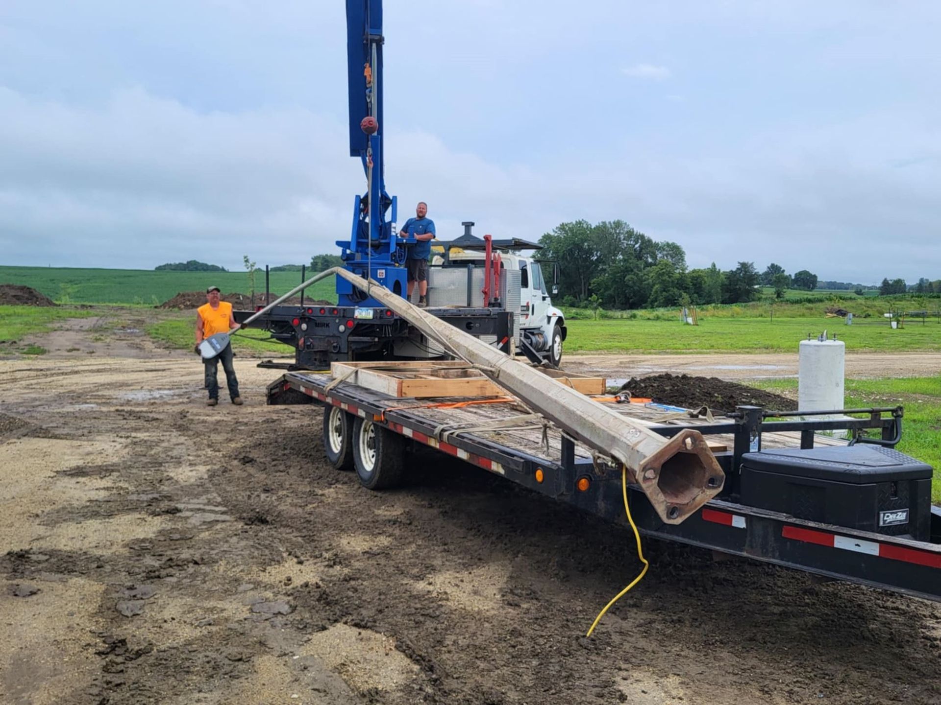 Workers loading long metal poles onto a truck at a muddy construction site with a drilling rig