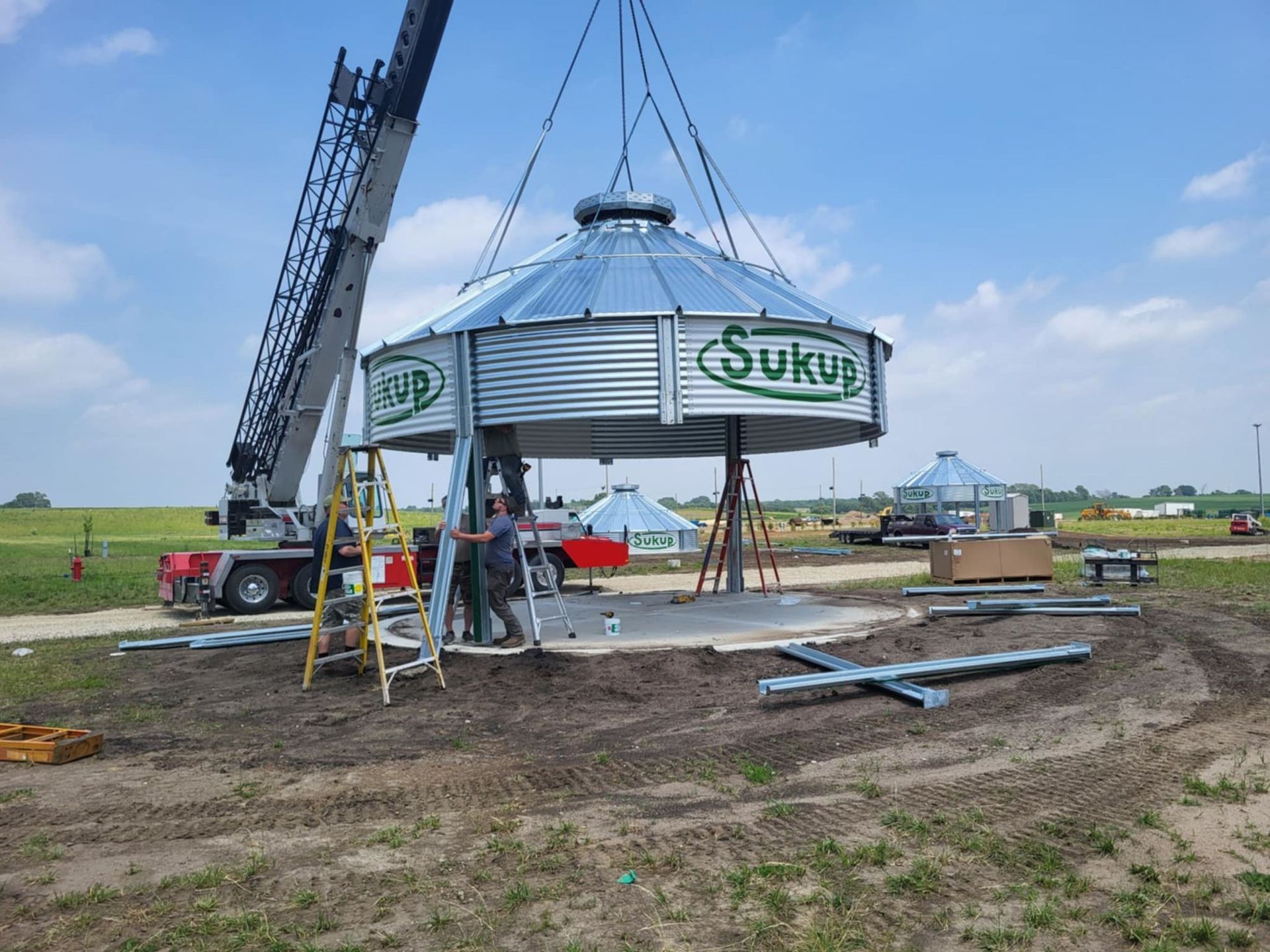 Workers install a large silver tank with a crane in a grassy field under a blue sky.